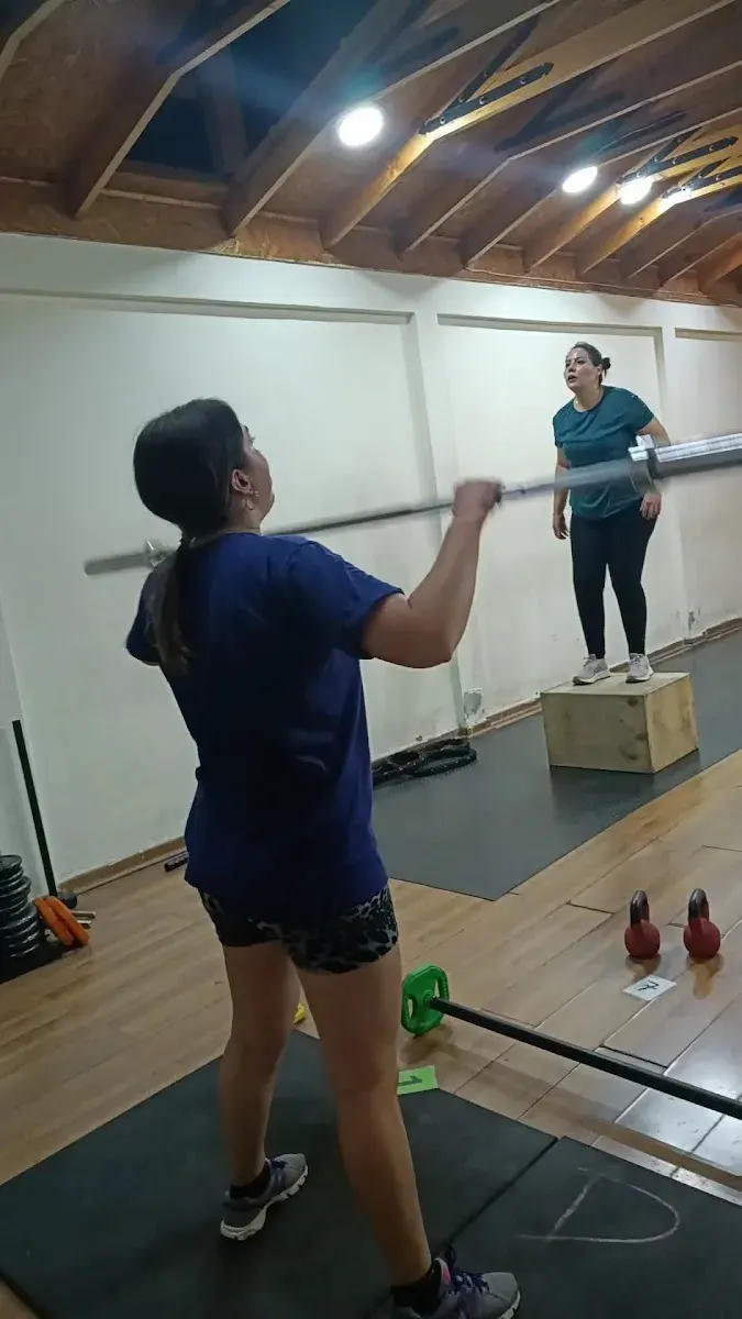 Alumna levantando barra mientras el instructor guía desde un cajón pliométrico