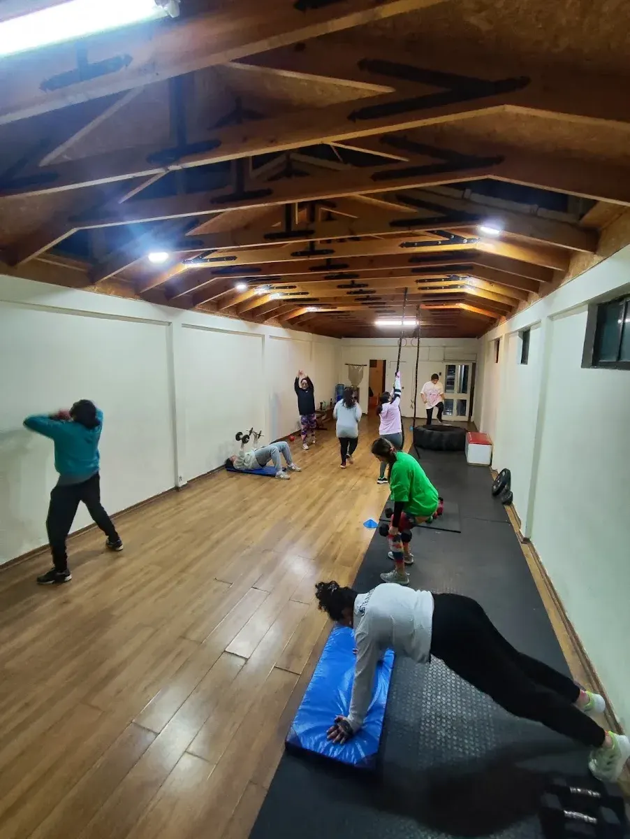 Clase en la sala de Atenas: alumnos entrenando con mancuernas, colchonetas y pliométricos bajo el techo de vigas de madera