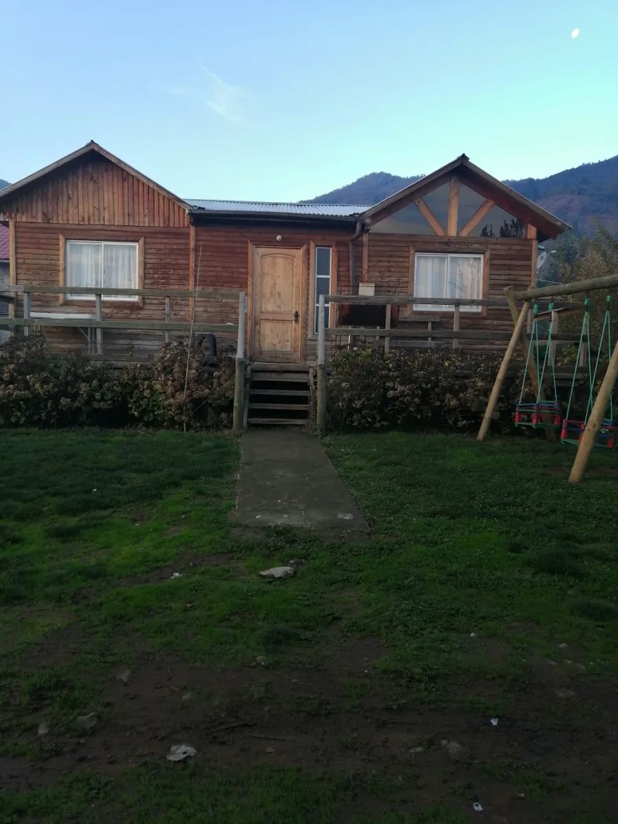 Cabañas El Alto desde el jardín: dos cabañas de madera sobre pradera verde con árboles