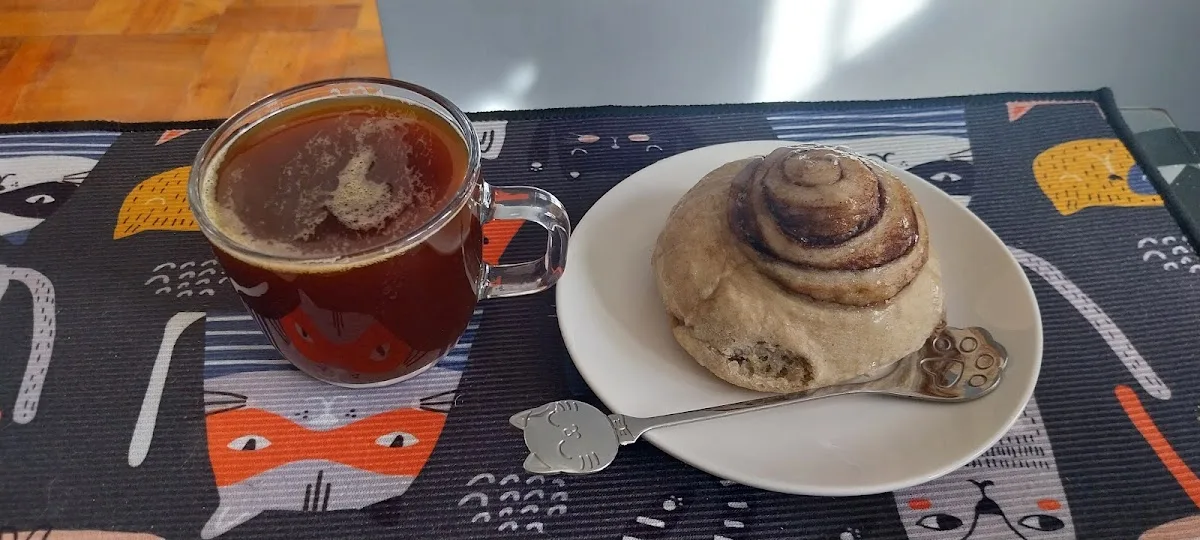 Té servido en taza blanca junto a un cinnamon roll, sobre el mantel con gatos de Catffeine