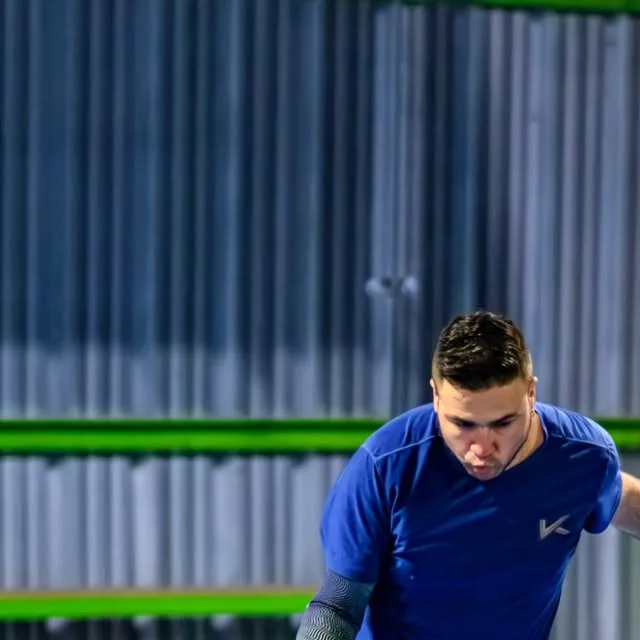 Jugador de polera azul preparando una devolución de derecha junto a la estructura verde lima