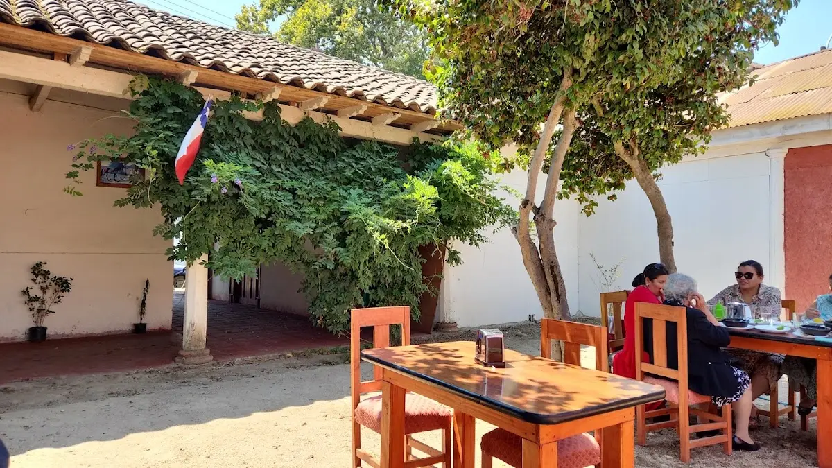 Patio del Club Social Yerbas Buenas: mesas con manteles bajo el parrón, la bandera chilena en el muro de adobe y clientes almorzando