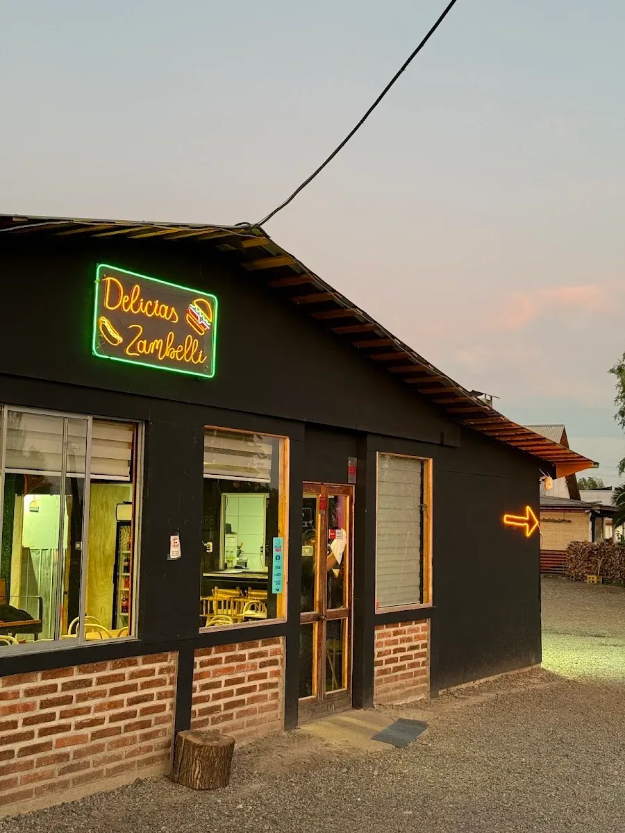 Fachada de Delicias Zambelli al anochecer, con el letrero de neón encendido y las ventanas iluminadas