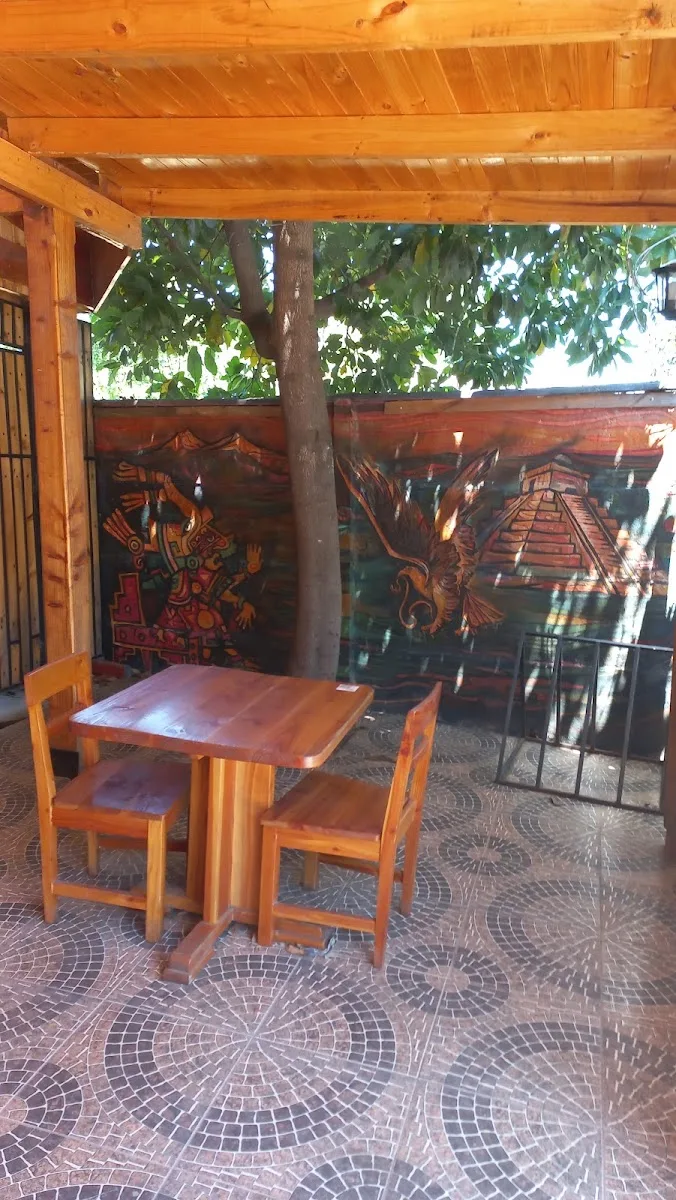 Terraza de madera con mesa bajo los árboles y mural prehispánico