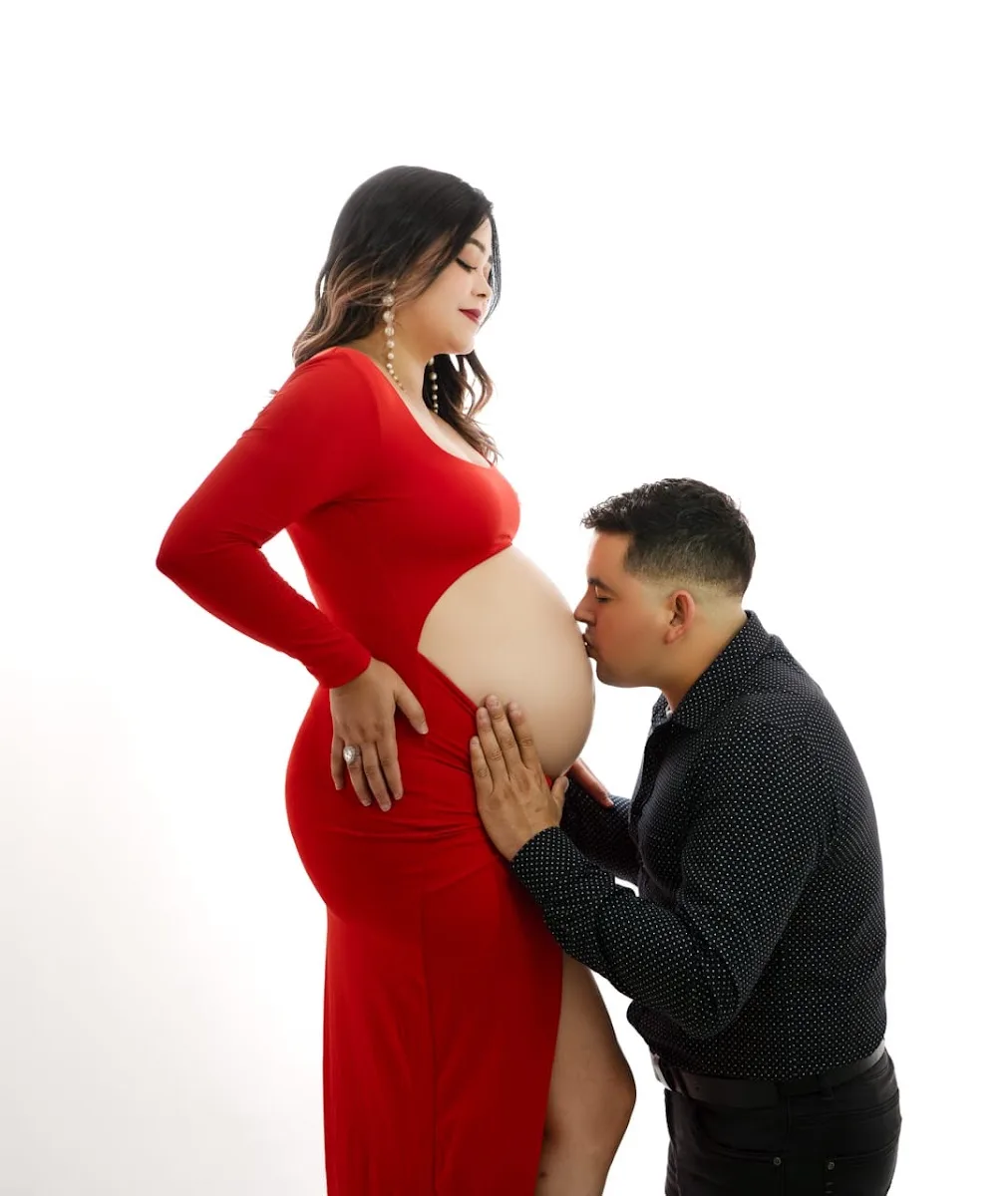 Embarazada con vestido rojo largo mientras su pareja besa la guatita, sobre fondo blanco de estudio