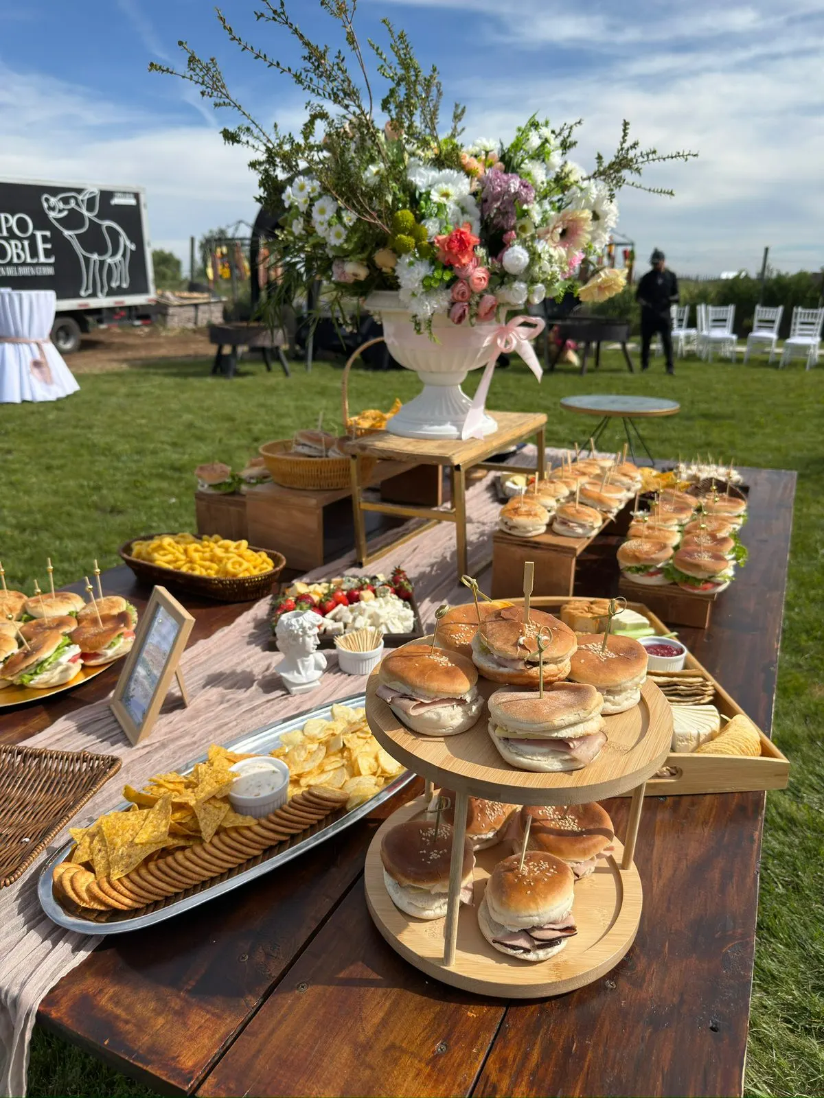 Mesa de cóctel con sándwiches, tablas de picoteo y arreglo floral en un evento de campo