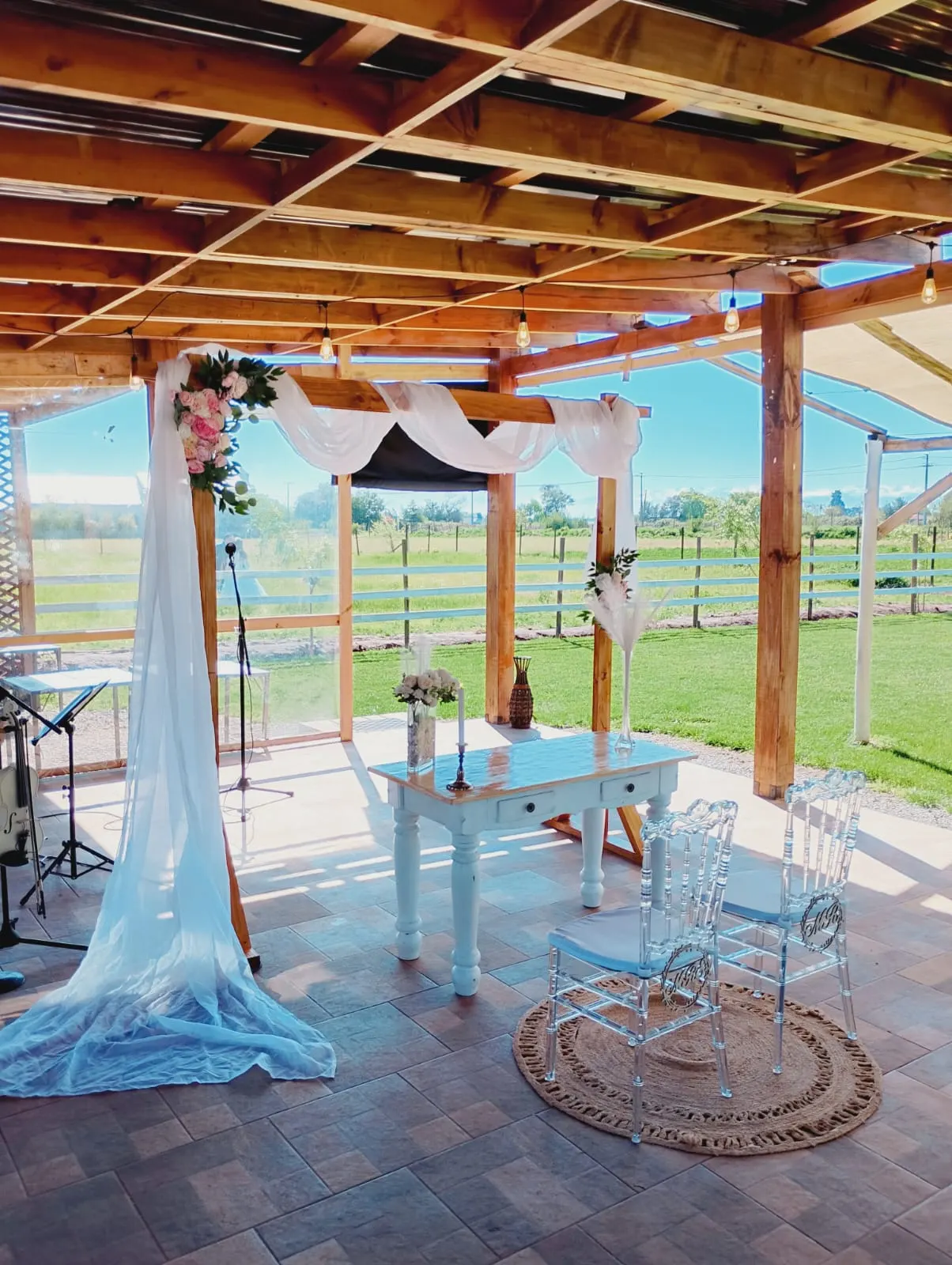 Pérgola decorada con telas y flores, sillas transparentes y mesa de ceremonia