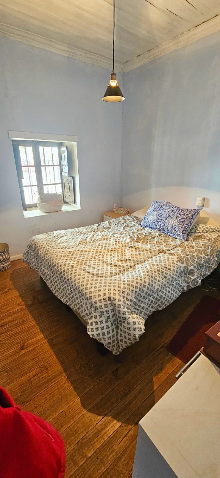Pieza del hospedaje: cama de dos plazas con cubrecama estampada, muro de adobe con ventana profunda y piso de madera