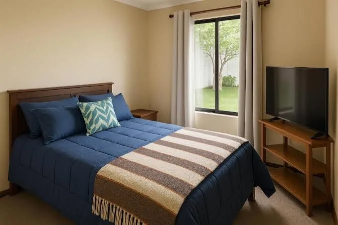 Habitación con cama azul, ventana con cortina y televisión en el muro