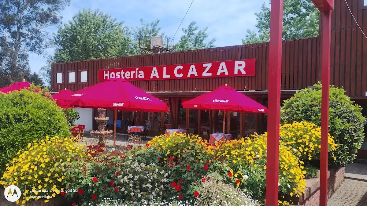 Fachada roja de Hostería Alcázar con quitasoles y jardín de flores, a un costado de la Ruta 5