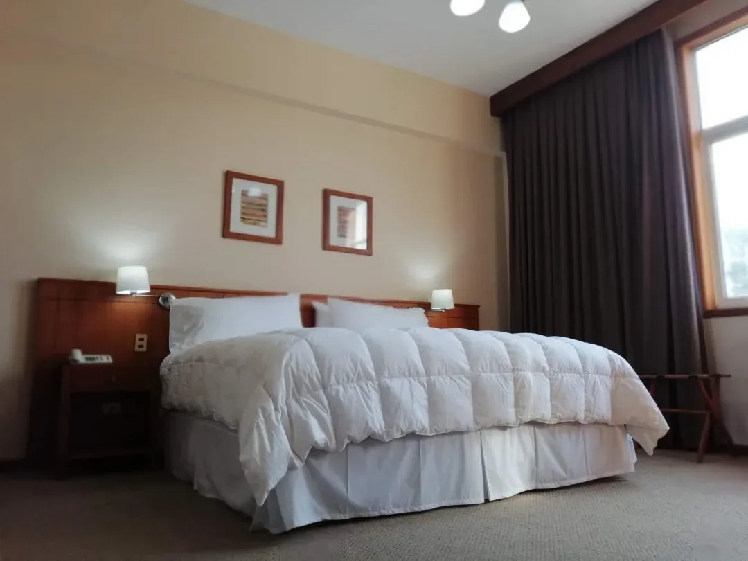Habitación doble con cama blanca, escritorio de madera y ventanal con cortinas
