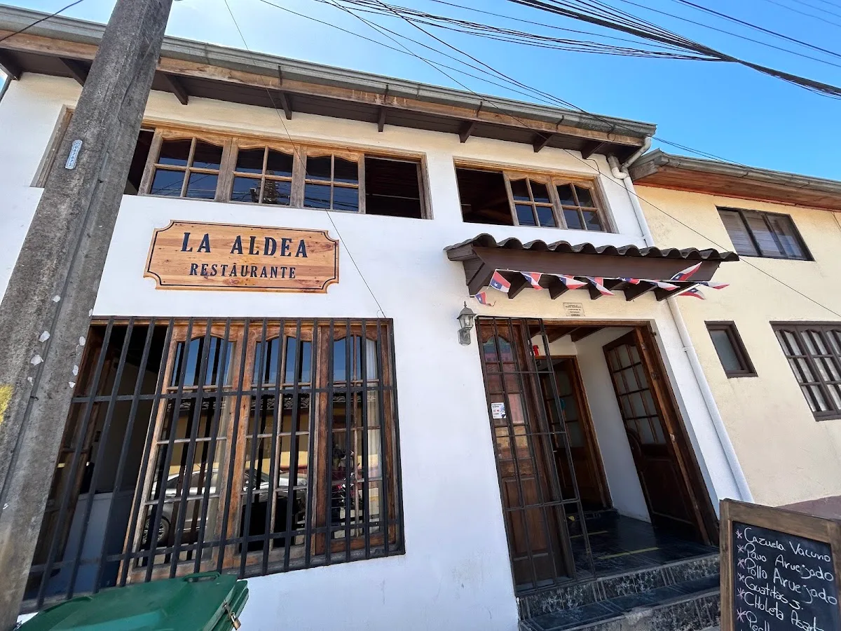 Fachada de La Aldea: casa blanca con letrero de madera tallado, banderines chilenos y pizarra en la puerta