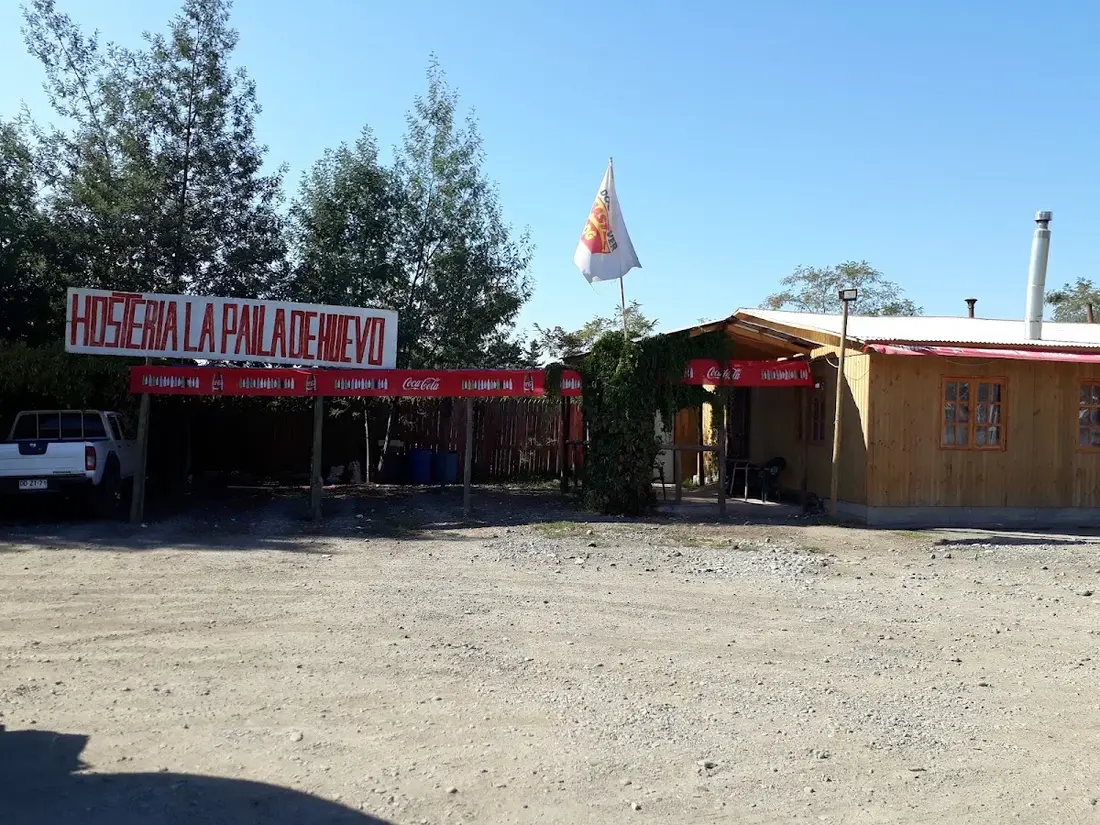 Fachada de la Hostería La Paila de Huevo: quincho de madera, letrero pintado y estacionamiento de ripio