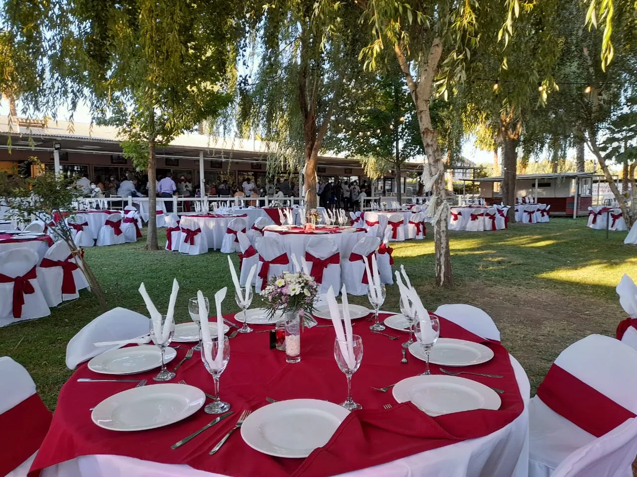 Mesas redondas con manteles rojos montadas en el prado, bajo los árboles, con el salón de Rancho Itahue al fondo
