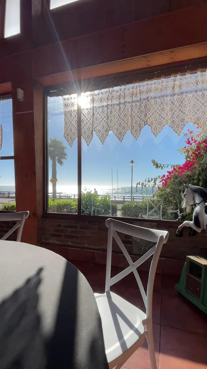 Mesa de Casa del Mar junto al ventanal: mantel blanco, cortina de crochet y el Pacífico al fondo