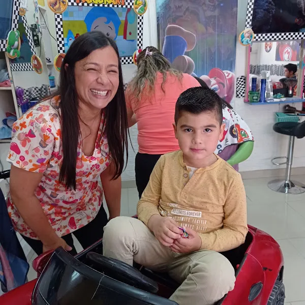 Niño feliz recién peinado en una silla-auto roja junto a su mamá y la estilista